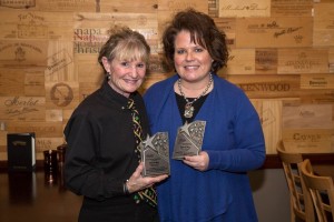 Two women standing together holding employee recognition awards in front of wood wall.