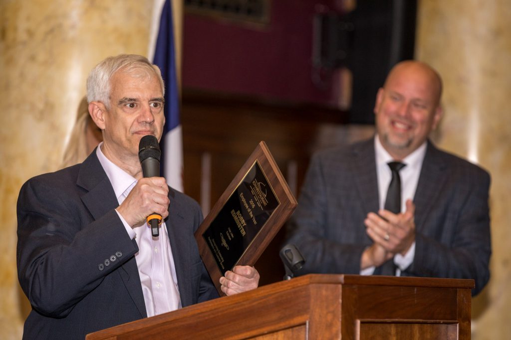 Man speaking into microphone holding award plaque while another man applauds in background.