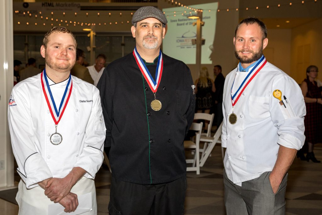 Three chefs wearing medals and uniforms posing together at culinary event.