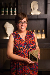 Woman with glasses in colorful red and blue patterned dress holding award trophy in bar or distillery setting.