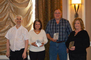 Four people holding awards and smiling at an indoor event with gold curtains.