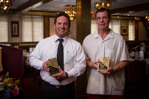 Two men holding awards and smiling in a restaurant setting with tables and chandeliers.