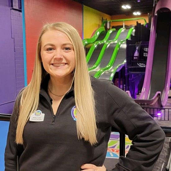 Woman in black jacket with name badge standing in front of colorful indoor playground slides.