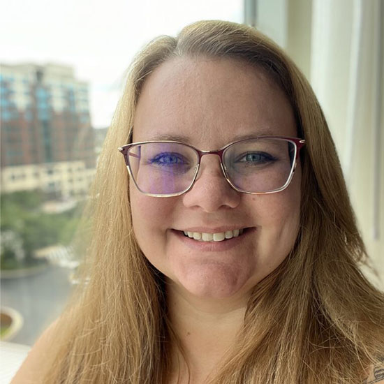 Woman with glasses and long light brown hair smiling by a window.