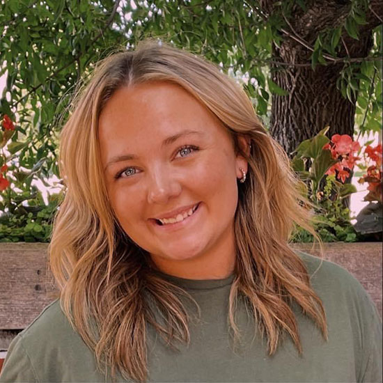 Woman with wavy blonde highlighted hair in olive green t-shirt smiling outdoors with trees in background.
