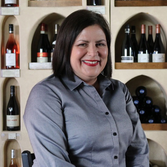 Woman with dark bob haircut in gray button-up shirt smiling in front of wine bottle display.