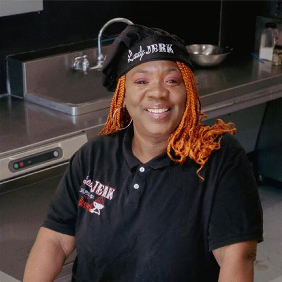 Smiling Black woman with orange braids wearing black chef visor and polo shirt in commercial kitchen.