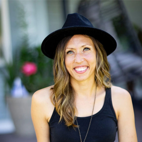 Woman with wavy blonde hair wearing black fedora hat and black tank top smiling outdoors.