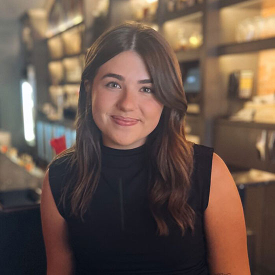Young woman with long dark brown hair in black sleeveless top in restaurant setting.