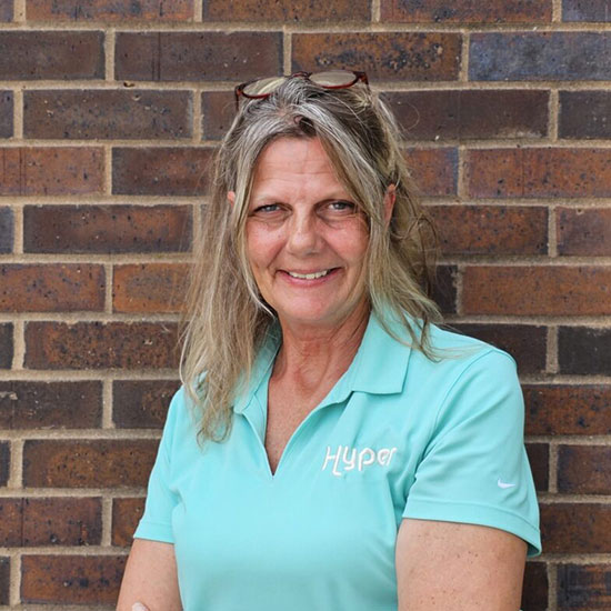 Woman with long gray-blonde hair and sunglasses on head in turquoise Hyper polo shirt against brick wall.