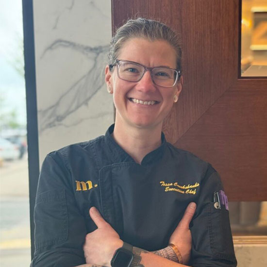 Executive chef in black chef jacket with glasses giving thumbs up in restaurant kitchen.