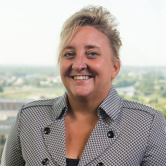 Professional woman with short blonde hair in patterned blazer smiling in office setting with city view.