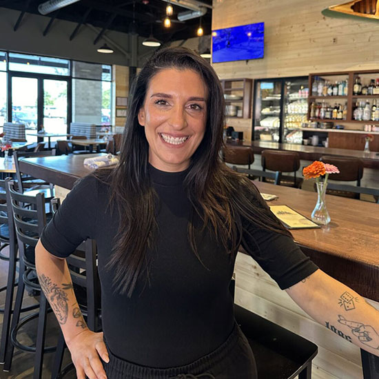 Smiling woman with long dark hair and arm tattoos standing in modern restaurant dining room.