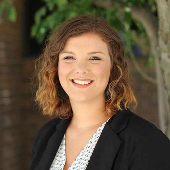 Woman with short curly hair wearing a black blazer and patterned top, smiling outdoors near a tree.