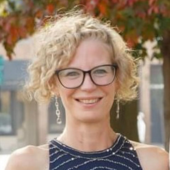 Blonde woman with curly hair and glasses wearing a sleeveless patterned dress, smiling outdoors with autumn trees in the background.