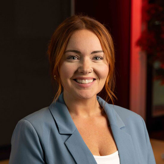 Woman with red hair in a blue blazer and white top, smiling warmly in a softly lit environment.