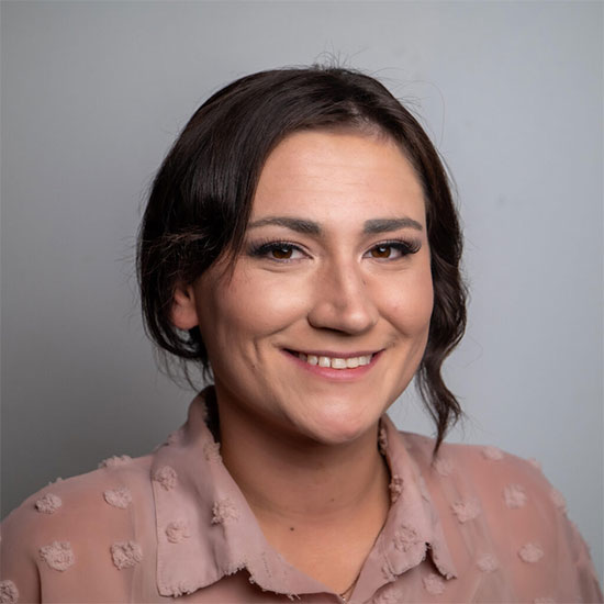 Close-up portrait of a woman with dark brown hair in a blush textured blouse, smiling in front of a neutral backdrop.