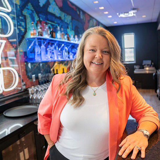 Woman in a coral blazer and white top, smiling behind a bar with a colorful mural background.