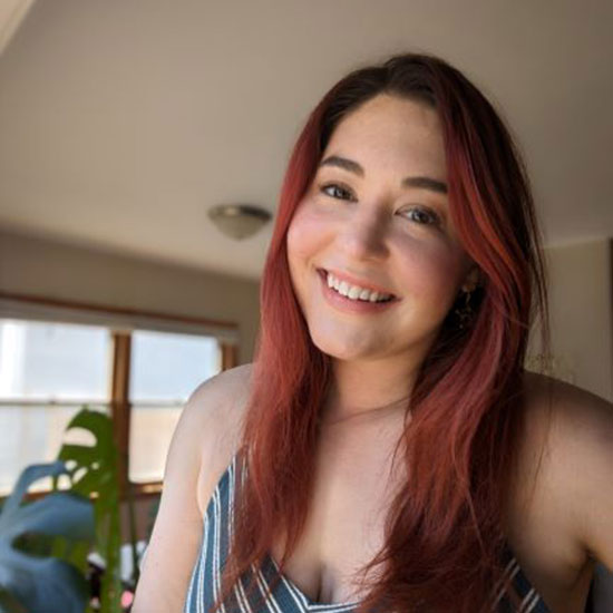 Woman with red hair wearing a striped dress, smiling in natural light with a plant visible in the background.