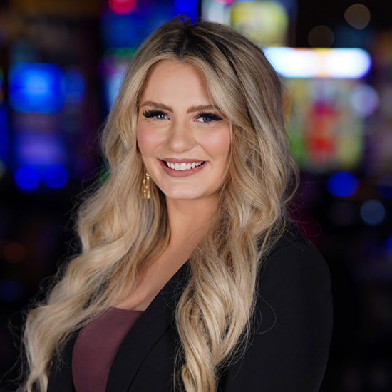 Woman with long blonde wavy hair in black blazer smiling with colorful bokeh lights in background.
