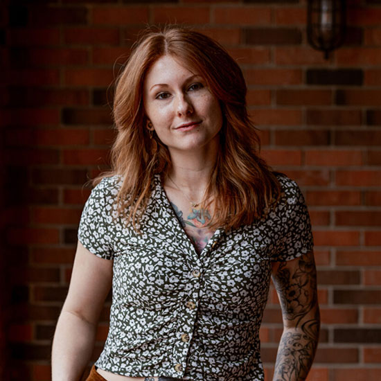 Woman with long red hair wearing floral print top and arm tattoos against brick wall.