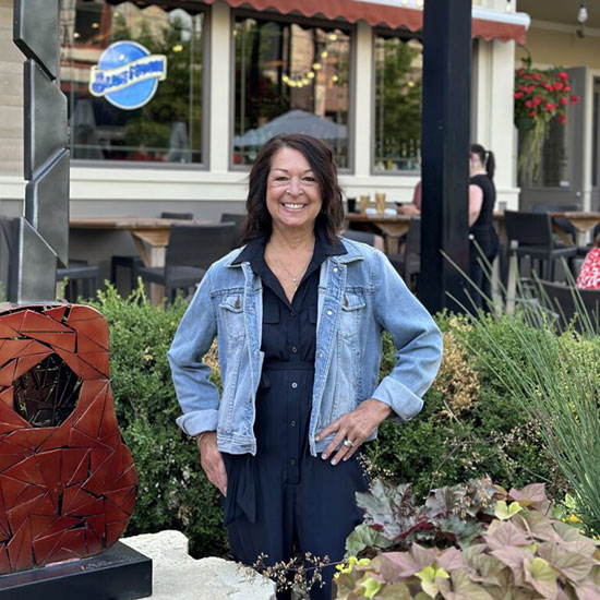 Woman in denim jacket smiling outside restaurant with decorative metal art and plants.