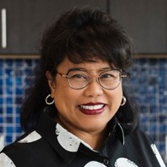 Professional woman wearing glasses and hoop earrings smiling in kitchen with blue tile backsplash.