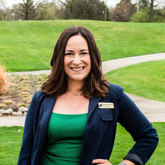 Woman in navy blazer and green top with name badge smiling in park setting.
