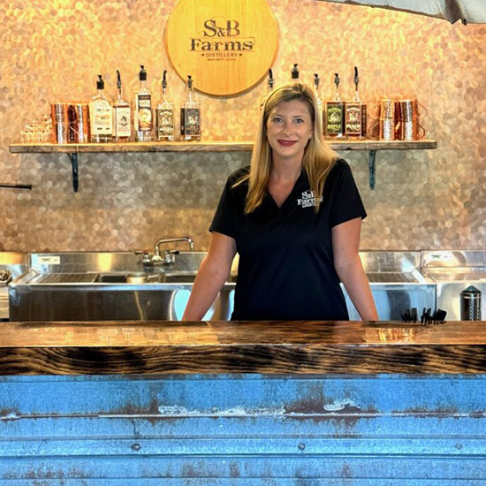 Woman standing behind the bar at S&B Farms Distillery with bottles displayed on shelves.
