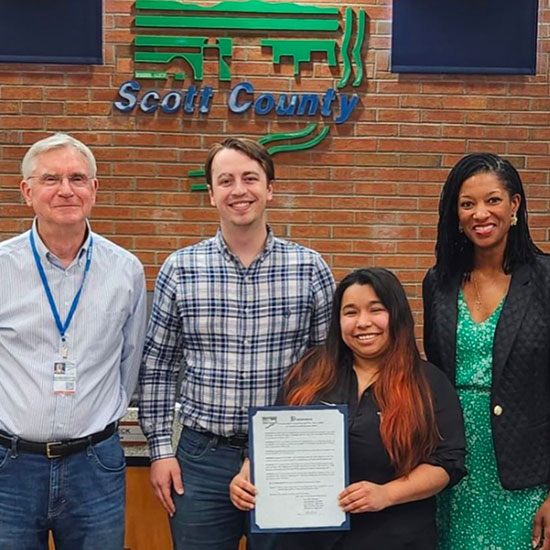 Group of four people standing in front of a Scott County sign holding an official document.