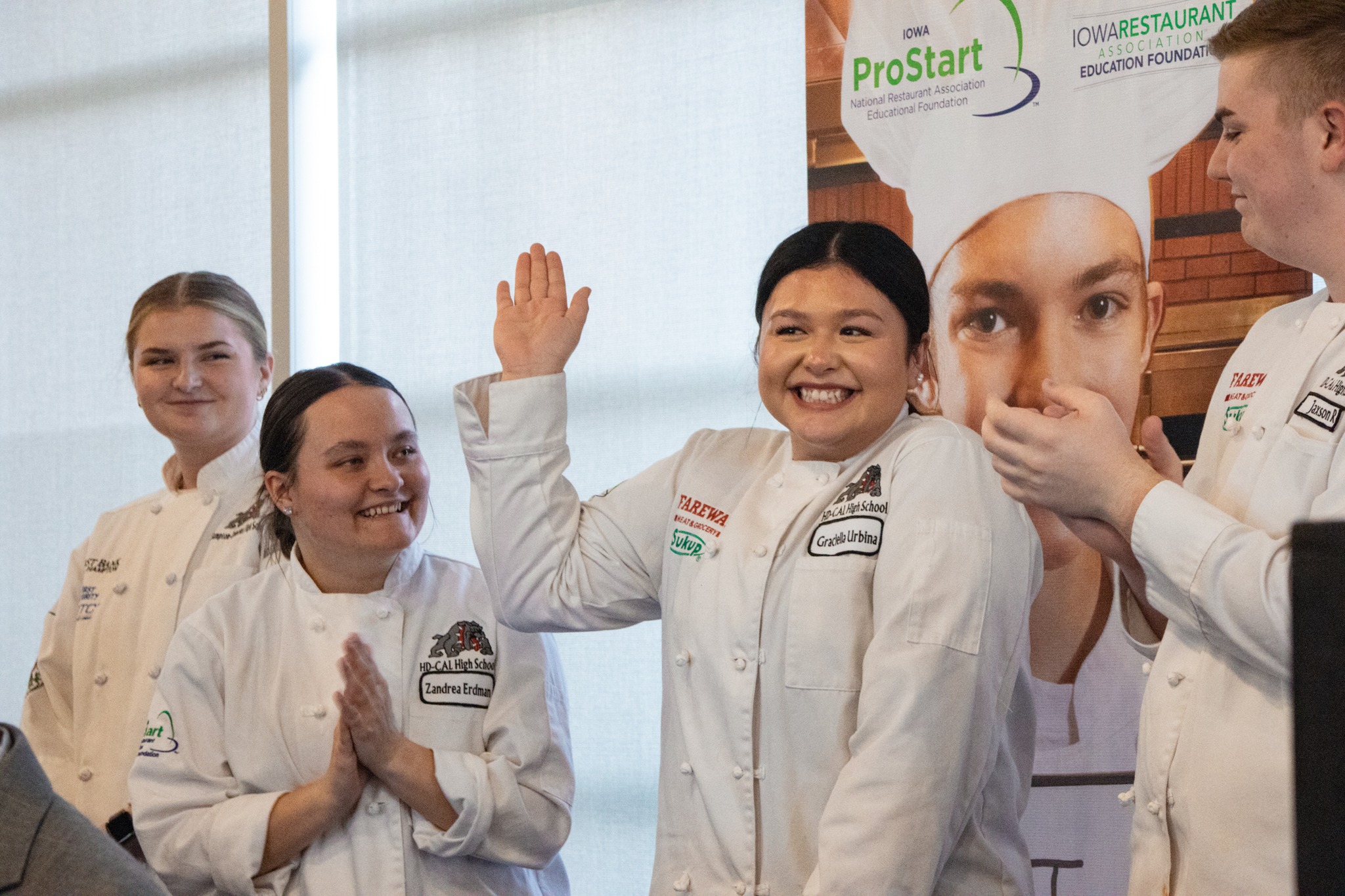 Culinary students in chef coats smiling on stage, one student raising her hand while others applaud.