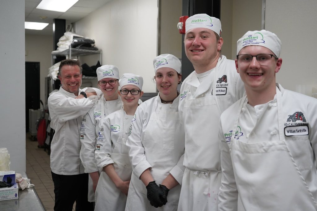 Culinary instructor standing with smiling ProStart students in chef uniforms.