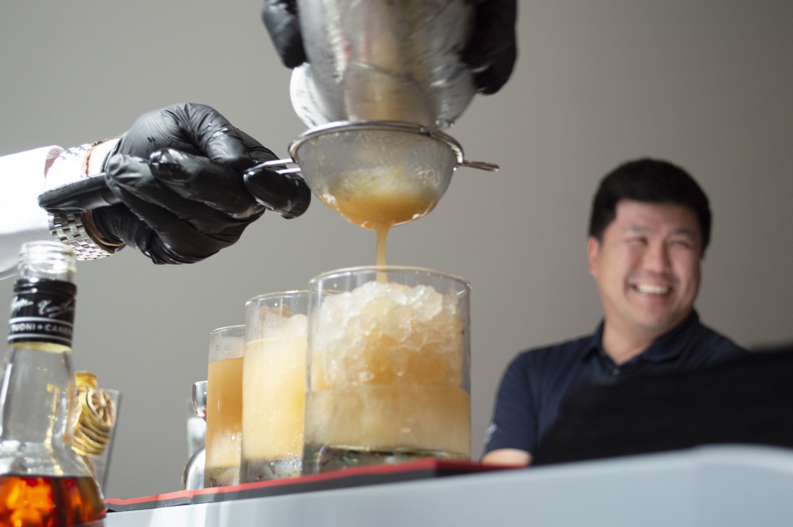 Bartender straining cocktail into glasses while attendee smiles in background.