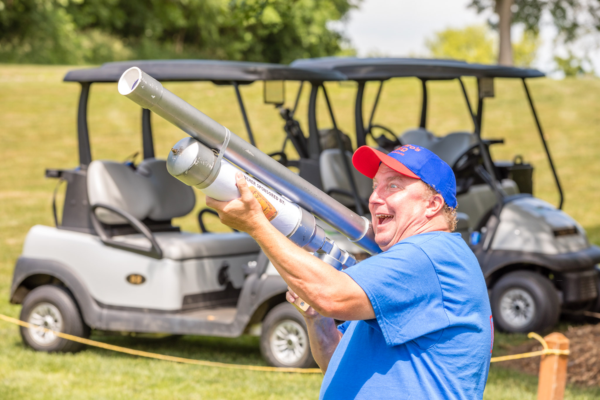 Golfer smiling while holding novelty launcher on course with carts in background.