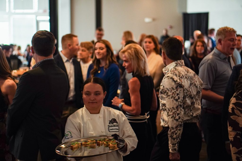 Culinary student in chef coat carrying a tray of appetizers at a crowded networking event.