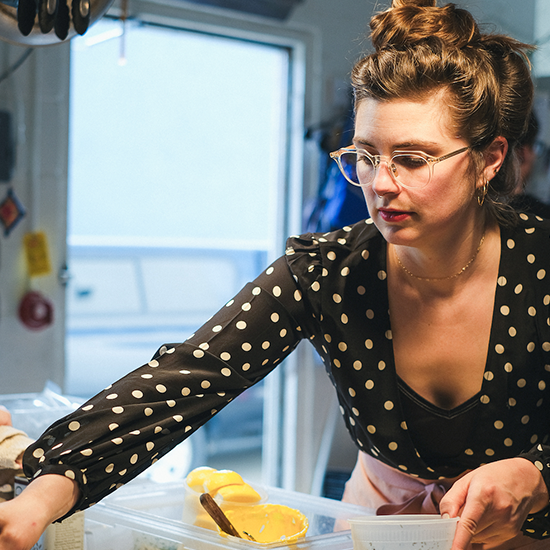 Woman with glasses and hair tied up, wearing a black polka-dot blouse, working with containers of food in a kitchen.