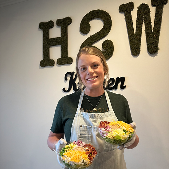 Smiling woman in an apron holding two bowls of salad, standing in front of a wall with “H2W Kitchen” signage.