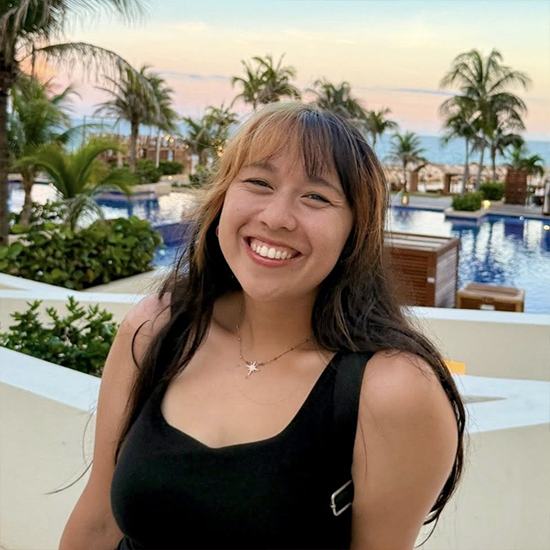 Smiling woman with long dark hair and light bangs, wearing a black dress, standing near a poolside resort at sunset.