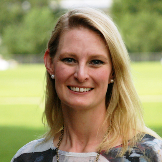 Outdoor headshot of a woman with blonde hair, pearl earrings, and a patterned top, smiling against a green backdrop.