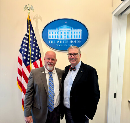 Two men in suits standing in front of the White House emblem and U.S. flag, smiling together.