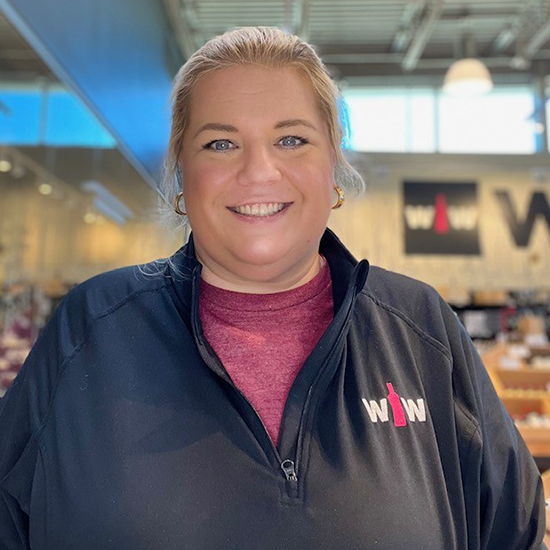 Smiling woman with blonde hair pulled back, wearing a black WOW-branded jacket, standing in a retail store.