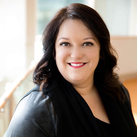 Professional headshot of a woman with dark brown hair, wearing a black outfit, smiling indoors with natural light.
