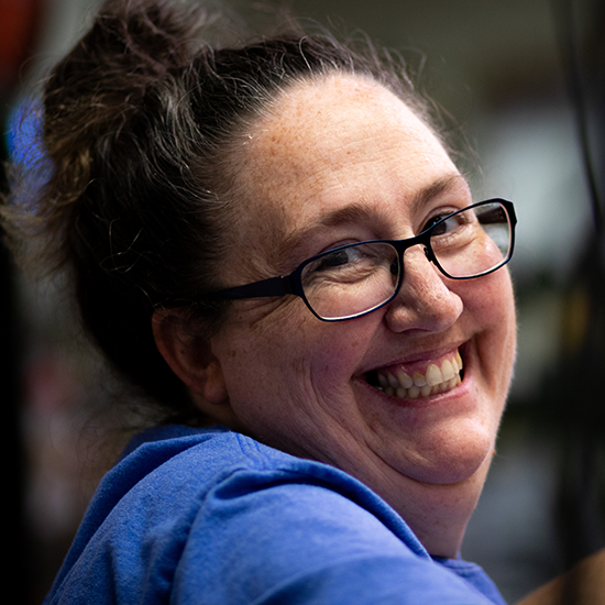 Smiling woman with glasses and tied-back hair, wearing a blue shirt in an indoor setting.
