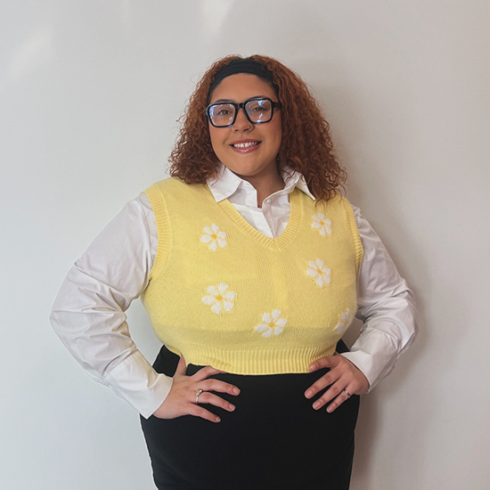 Woman with curly red hair wearing glasses, a yellow sweater vest with white flowers, and a white collared shirt.