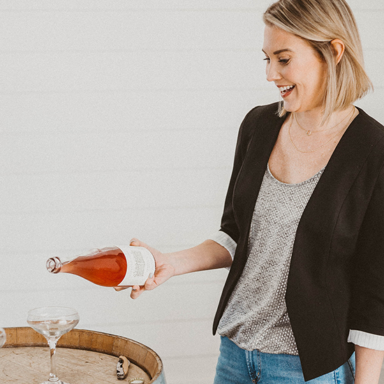 Woman with blonde hair wearing a black blazer and gray top, pouring rosé wine into a glass from a bottle.