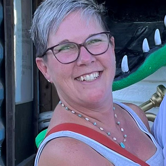 Woman with short gray hair, glasses, and a necklace smiling outdoors in casual clothing.