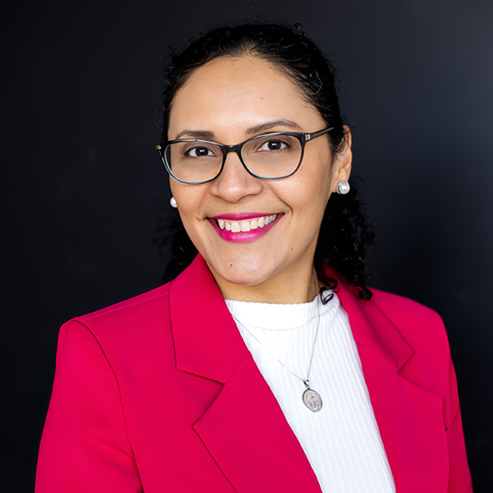 Professional headshot of a smiling woman with glasses wearing a bright pink blazer over a white top and pendant necklace against a dark background.