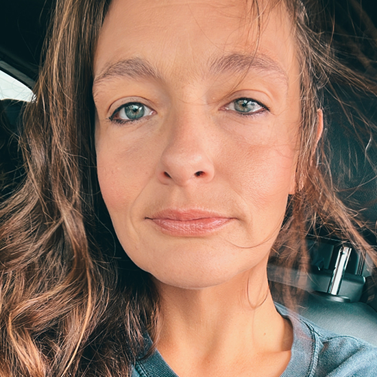 Close-up portrait of a woman with long brown hair and blue eyes, wearing a casual top in a car.