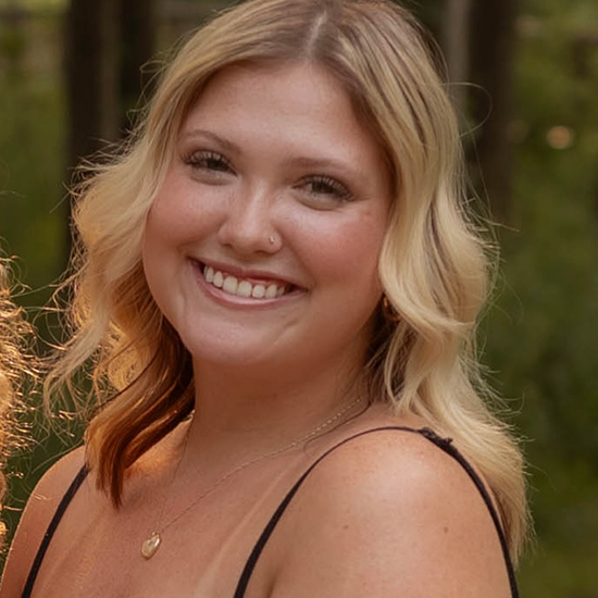 Smiling woman with wavy blonde hair, wearing a necklace and black top, photographed outdoors.