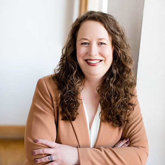 Professional headshot of a woman with long curly hair, wearing a tan blazer and white blouse, smiling with arms crossed.